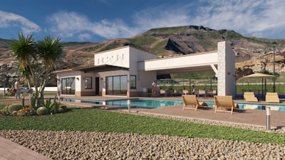 A modern house is situated in a scenic mountainous backdrop. The structure is a single-story building with large windows and stone accents. In the foreground, there is a swimming pool surrounded by a patio with wooden lounge chairs. The landscape features well-manicured grass, desert plants, and a section of decorative gravel.