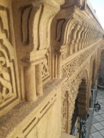 Intricate carvings on the façade of a building with a series of arches, displaying detailed patterns and designs. The architecture shows a high level of craftsmanship. A tiled floor and some hanging lamps can be seen below the structure.