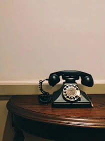 Close-up of a classic rotary phone resting on a polished wooden table with a vintage wallpaper backdrop.