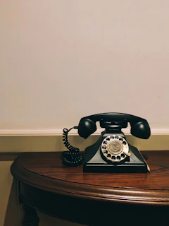 Close-up of a classic rotary phone resting on a polished wooden table with a vintage wallpaper backdrop.