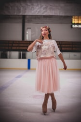 A person on an ice rink is wearing figure skates and a pink tulle skirt paired with a white t-shirt featuring an image or print. A flower crown adorns the head, adding a whimsical touch. The background includes ice rink barriers and a scoreboard with visible but indistinct numbers.
