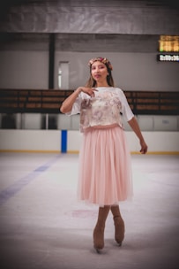 A person on an ice rink is wearing figure skates and a pink tulle skirt paired with a white t-shirt featuring an image or print. A flower crown adorns the head, adding a whimsical touch. The background includes ice rink barriers and a scoreboard with visible but indistinct numbers.