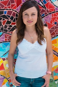 A person with long brown hair, wearing a white sleeveless top and jeans, stands in front of a vibrant mosaic wall. The mosaic features an array of geometric patterns in various colors, including red, blue, purple, and yellow.
