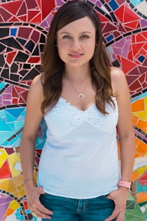 A person with long brown hair, wearing a white sleeveless top and jeans, stands in front of a vibrant mosaic wall. The mosaic features an array of geometric patterns in various colors, including red, blue, purple, and yellow.