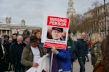 A group of people are gathered outdoors in a public square. One person is holding a large sign with the heading 'MISSING PERSON' in bold red letters and an image of a man below it. The sign includes text about Jeremy Corbyn. The background features historic buildings and a clock tower, with some people looking engaged and others walking around.