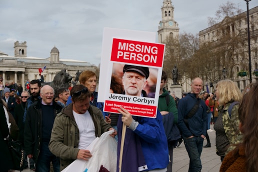 A group of people are gathered outdoors in a public square. One person is holding a large sign with the heading 'MISSING PERSON' in bold red letters and an image of a man below it. The sign includes text about Jeremy Corbyn. The background features historic buildings and a clock tower, with some people looking engaged and others walking around.