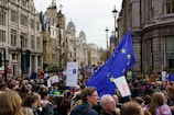 A large crowd is gathered on a city street, participating in a protest or demonstration. The crowd is dense, with people holding signs and flags. A prominent European Union flag is visible, indicating the theme of the protest. The street is lined with historic architecture, and the day appears overcast.