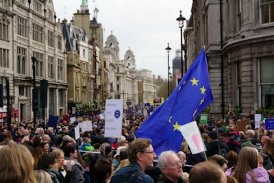 A large crowd is gathered on a city street, participating in a protest or demonstration. The crowd is dense, with people holding signs and flags. A prominent European Union flag is visible, indicating the theme of the protest. The street is lined with historic architecture, and the day appears overcast.