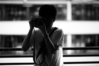 A silhouette of a person holding a camera to their face, standing in front of a railing. The background appears to be an out-of-focus urban environment with a modern building featuring large windows. The high contrast between light and shadow creates a dramatic effect.