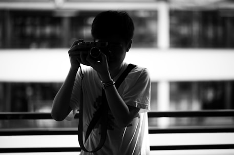 A silhouette of a person holding a camera to their face, standing in front of a railing. The background appears to be an out-of-focus urban environment with a modern building featuring large windows. The high contrast between light and shadow creates a dramatic effect.