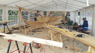 A wooden boat under construction inside a large white tent, surrounded by various tools and lumber. Three men are present, working on the boat or seated nearby. The environment is well-lit with natural light filtering through the tent windows, and the floor is covered in wood shavings.