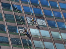 Two window cleaners are suspended by ropes, working on cleaning the glass windows of a tall building. Each cleaner is equipped with tools and a bucket, skillfully moving along the facade with safety harnesses. The building features reflective blue-tinted glass panels, creating a striking geometric pattern.