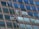 Two window cleaners are suspended by ropes, working on cleaning the glass windows of a tall building. Each cleaner is equipped with tools and a bucket, skillfully moving along the facade with safety harnesses. The building features reflective blue-tinted glass panels, creating a striking geometric pattern.