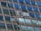 Two window cleaners are suspended by ropes, working on cleaning the glass windows of a tall building. Each cleaner is equipped with tools and a bucket, skillfully moving along the facade with safety harnesses. The building features reflective blue-tinted glass panels, creating a striking geometric pattern.