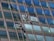 Two window cleaners are suspended by ropes, working on cleaning the glass windows of a tall building. Each cleaner is equipped with tools and a bucket, skillfully moving along the facade with safety harnesses. The building features reflective blue-tinted glass panels, creating a striking geometric pattern.