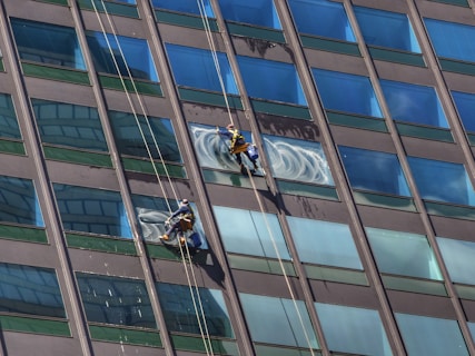 Two window cleaners are suspended by ropes, working on cleaning the glass windows of a tall building. Each cleaner is equipped with tools and a bucket, skillfully moving along the facade with safety harnesses. The building features reflective blue-tinted glass panels, creating a striking geometric pattern.