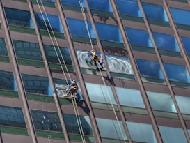 Two window cleaners are suspended by ropes, working on cleaning the glass windows of a tall building. Each cleaner is equipped with tools and a bucket, skillfully moving along the facade with safety harnesses. The building features reflective blue-tinted glass panels, creating a striking geometric pattern.