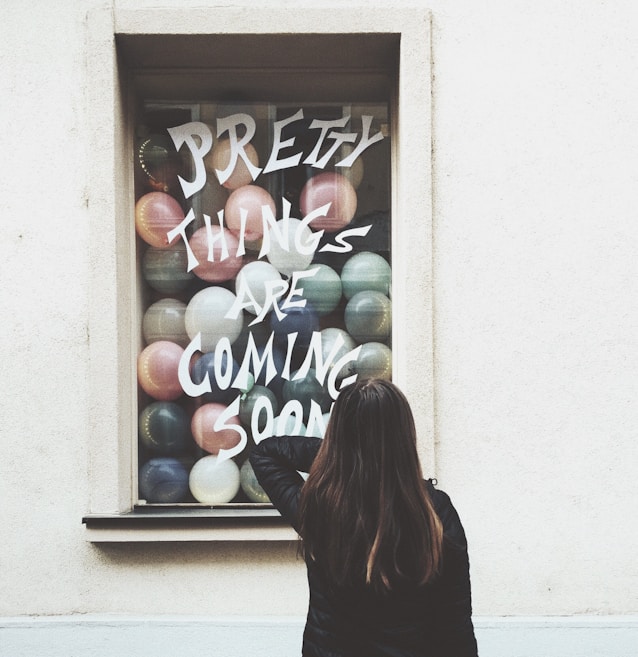 A person with long hair is standing in front of a window display filled with colorful balloons. The window has the phrase 'Pretty Things Are Coming Soon' written on it in bold, artistic lettering.