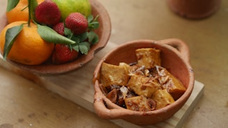 A rustic terracotta bowl filled with fried tofu cubes, garnished with caramelized onions. Next to it is a clay dish containing an arrangement of fresh fruits including oranges, strawberries, and a pear, with green leaves for decoration, all placed on a wooden surface.