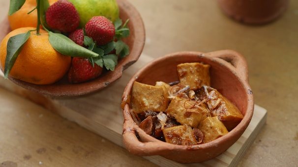 A rustic terracotta bowl filled with fried tofu cubes, garnished with caramelized onions. Next to it is a clay dish containing an arrangement of fresh fruits including oranges, strawberries, and a pear, with green leaves for decoration, all placed on a wooden surface.