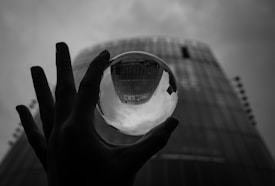 A monochrome image featuring a hand holding a small crystal ball, reflecting an urban skyline with a tall building. The background shows parts of a skyscraper blurred due to depth of field. The atmosphere looks moody with overcast skies.