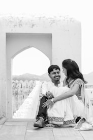 A couple is seated together in a charming outdoor setting, dressed in traditional attire. The man and woman are closely seated, sharing an affectionate gaze and smile. The background features an architectural archway and distant hills, providing a serene and romantic ambience.