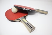 Two table tennis paddles with red rubber surfaces are placed on a white surface. A white ping pong ball is positioned between them, partially resting on one of the paddles. The paddles feature wooden handles with branding labels visible.