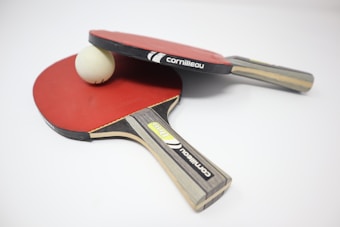 Two table tennis paddles with red rubber surfaces are placed on a white surface. A white ping pong ball is positioned between them, partially resting on one of the paddles. The paddles feature wooden handles with branding labels visible.