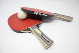 Two table tennis paddles with red rubber surfaces are placed on a white surface. A white ping pong ball is positioned between them, partially resting on one of the paddles. The paddles feature wooden handles with branding labels visible.