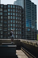 A person with a backpack and a cap stands on a rooftop ledge, taking a photograph with a camera. The background features modern high-rise buildings with multiple windows, including a round building and a glass facade. The scene is captured in daylight, with shadows creating a moody atmosphere.