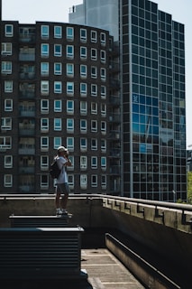 A person with a backpack and a cap stands on a rooftop ledge, taking a photograph with a camera. The background features modern high-rise buildings with multiple windows, including a round building and a glass facade. The scene is captured in daylight, with shadows creating a moody atmosphere.