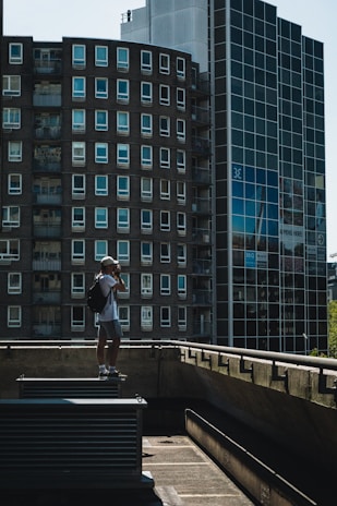 A person with a backpack and a cap stands on a rooftop ledge, taking a photograph with a camera. The background features modern high-rise buildings with multiple windows, including a round building and a glass facade. The scene is captured in daylight, with shadows creating a moody atmosphere.
