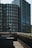 A person with a backpack and a cap stands on a rooftop ledge, taking a photograph with a camera. The background features modern high-rise buildings with multiple windows, including a round building and a glass facade. The scene is captured in daylight, with shadows creating a moody atmosphere.
