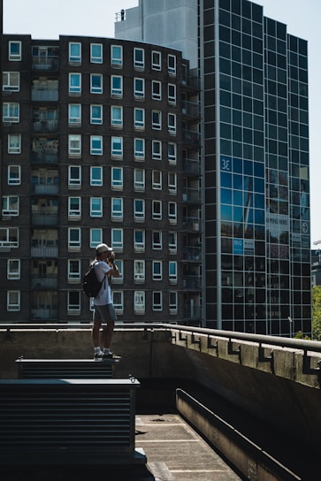 A person with a backpack and a cap stands on a rooftop ledge, taking a photograph with a camera. The background features modern high-rise buildings with multiple windows, including a round building and a glass facade. The scene is captured in daylight, with shadows creating a moody atmosphere.