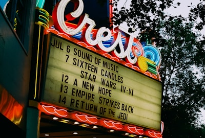 A vintage theater marquee displays a list of movie titles with showdates in bold black letters against a lighted background. Above the marquee, neon lights spell out 'Crest' in swirling, colorful designs, with surrounding decorative lights casting a nostalgic glow. Trees can be seen in the background against a dusky sky.