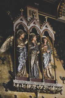 Three intricately carved and adorned statues of angels stand within ornate Gothic arches. Each figure is dressed in richly detailed garments with gilded accents, and they have halo-like decorations above their heads. The background features a decorative pattern, emphasizing the sacred and historical essence of the scene.