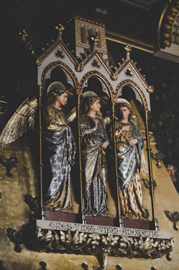 Three intricately carved and adorned statues of angels stand within ornate Gothic arches. Each figure is dressed in richly detailed garments with gilded accents, and they have halo-like decorations above their heads. The background features a decorative pattern, emphasizing the sacred and historical essence of the scene.