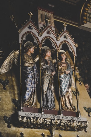 Three intricately carved and adorned statues of angels stand within ornate Gothic arches. Each figure is dressed in richly detailed garments with gilded accents, and they have halo-like decorations above their heads. The background features a decorative pattern, emphasizing the sacred and historical essence of the scene.