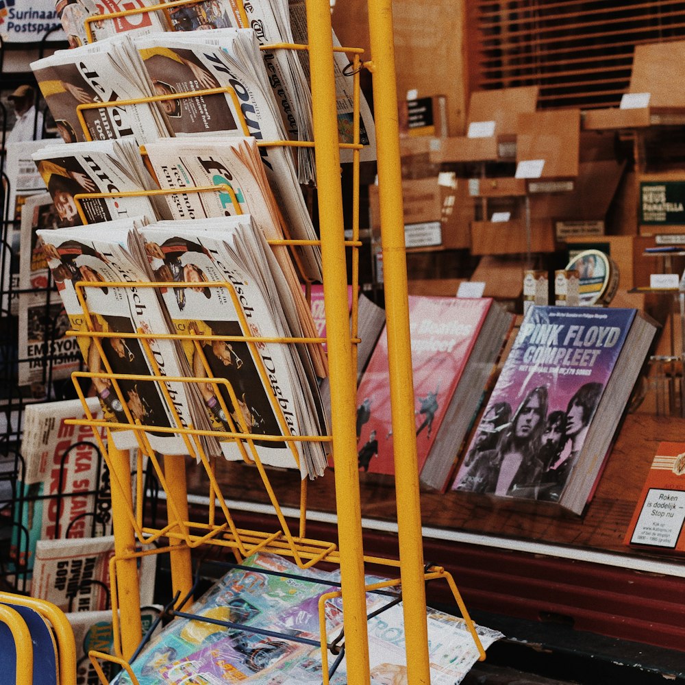Revistas e jornais variados lotam em rack de metal amarelo