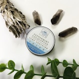 A tin of peppermint eucalyptus hemp soothing salve is placed on a white surface. Surrounding it are three dark crystals, a bundle of dried sage tied with string, and a sprig of green leaves.