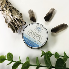 A tin of peppermint eucalyptus hemp soothing salve is placed on a white surface. Surrounding it are three dark crystals, a bundle of dried sage tied with string, and a sprig of green leaves.