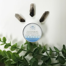 A circular container labeled 'Peppermint Eucalyptus Hemp Soothing Salve' is placed on a light background. Around it are three quartz crystals and green leafy stems.