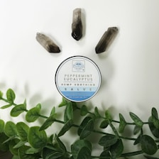 A circular container labeled 'Peppermint Eucalyptus Hemp Soothing Salve' is placed on a light background. Around it are three quartz crystals and green leafy stems.