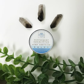 A circular container labeled 'Peppermint Eucalyptus Hemp Soothing Salve' is placed on a light background. Around it are three quartz crystals and green leafy stems.
