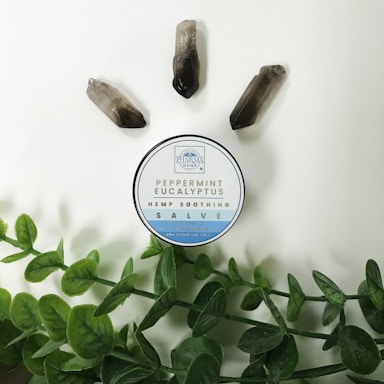 A circular container labeled 'Peppermint Eucalyptus Hemp Soothing Salve' is placed on a light background. Around it are three quartz crystals and green leafy stems.