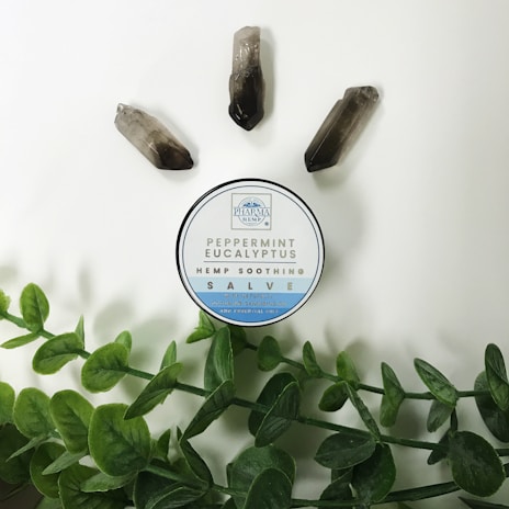A circular container labeled 'Peppermint Eucalyptus Hemp Soothing Salve' is placed on a light background. Around it are three quartz crystals and green leafy stems.