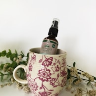 A floral-patterned ceramic mug holds a bottle labeled 'Pharma Hemp.' The mug features red and pink flowers against a white background. Surrounding the mug are sprigs of delicate white flowers with green leaves, adding a natural and calming touch.