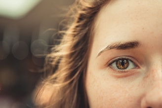 A close-up of a person's eye with a focus on the iris and surrounding skin. The eye is framed by light brown hair and the background is softly blurred. Freckles are visible on the skin, adding texture to the image.