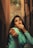 A woman with long dark hair and red lipstick poses in a narrow alleyway. She is wearing a teal sweater and extends her hand toward the camera with a thoughtful expression. The background is softly blurred, featuring warm tones from the walls.