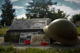 A gravestone with an inscription for a military individual is accompanied by a vintage helmet, black and white photographs, and two red poppies. Trees and a cloudy sky form the background, creating a serene and reflective setting.